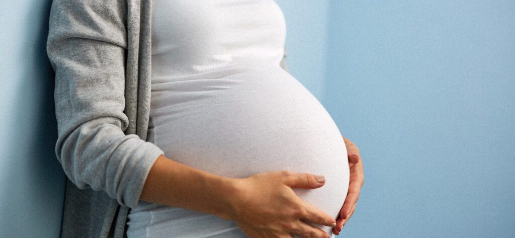 Woman during last months of pregnancy holding her belly standing against wall in blue room