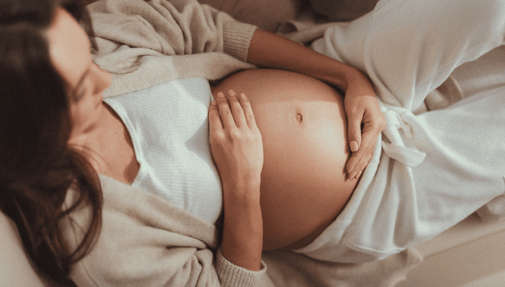 Pregnant woman in comfortable clothing lying down, holding her hands and looking at her pregnant belly.